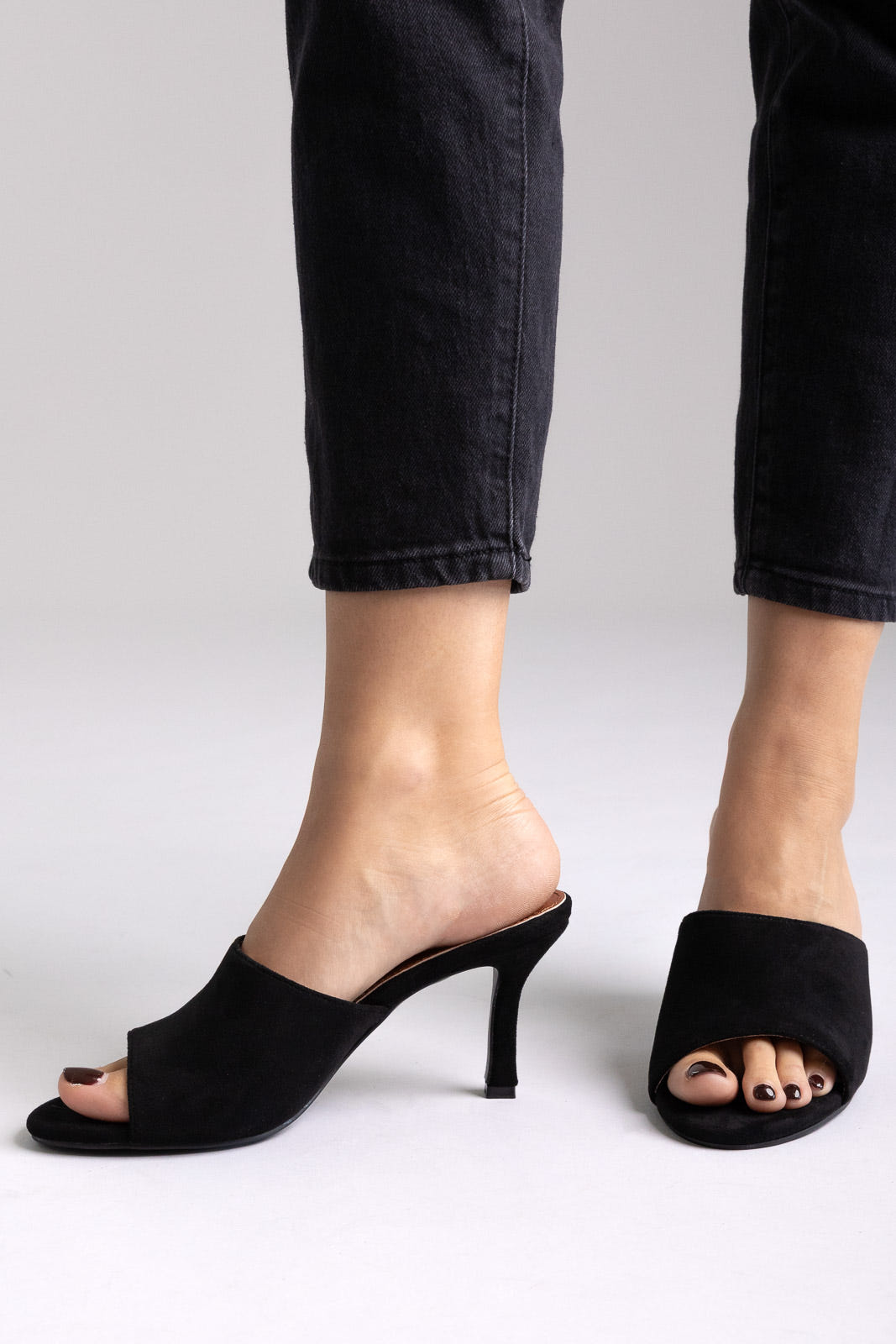 Black suede open-toe kitten-heel mules, side view on a model in black jeans in studio light