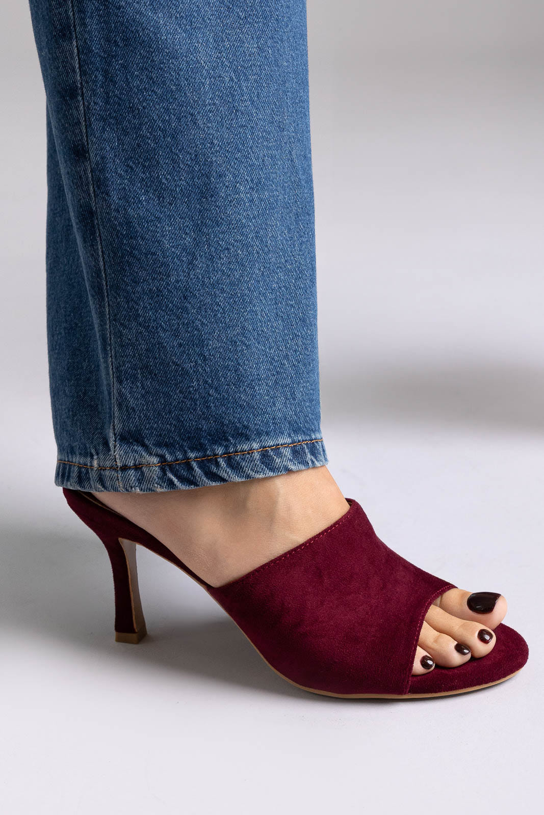 Burgundy suede open-toe mules with stiletto heel, angled view on a model in jeans