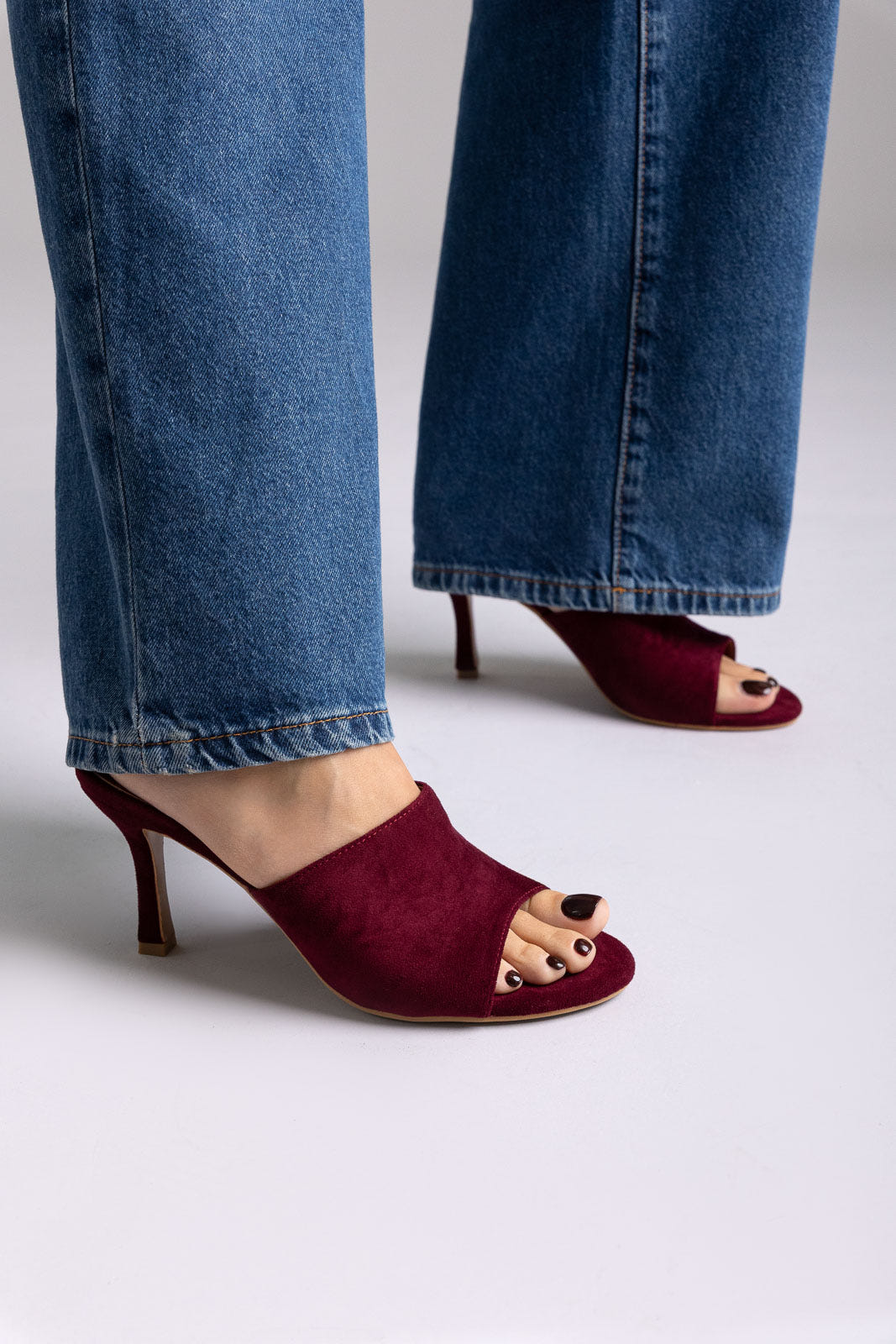 Burgundy suede open-toe mules with stiletto heel, angled view on a model in jeans