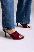 Burgundy suede open-toe mules with stiletto heel, angled view on a model in jeans