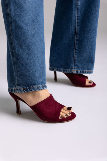 Burgundy suede open-toe mules with stiletto heel, angled view on a model in jeans