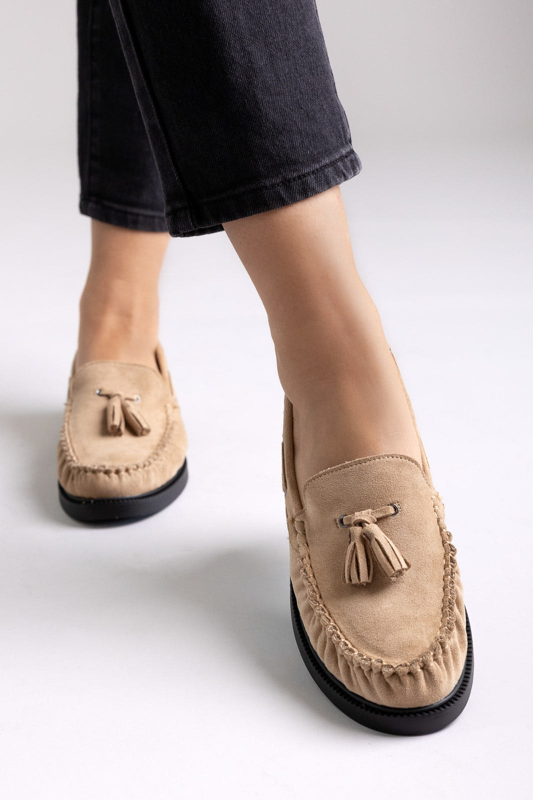 Taupe suede tassel loafers with rubber sole, angled view on a model in black jeans