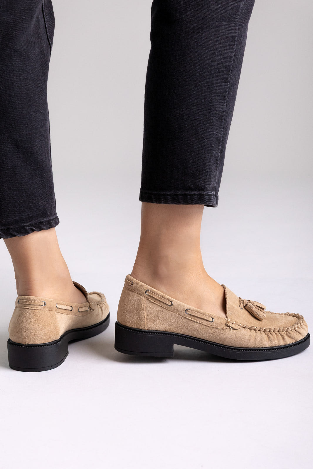 Taupe suede tassel loafers with rubber sole, angled view on a model in black jeans