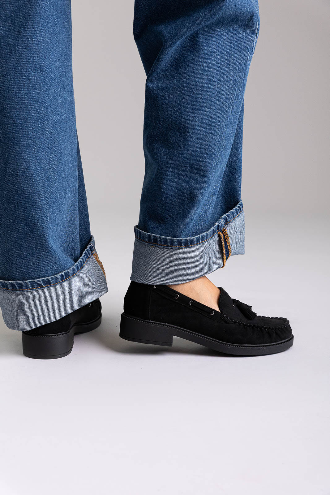 Black suede tassel loafers with flat sole, side view on a model in wide-leg jeans
