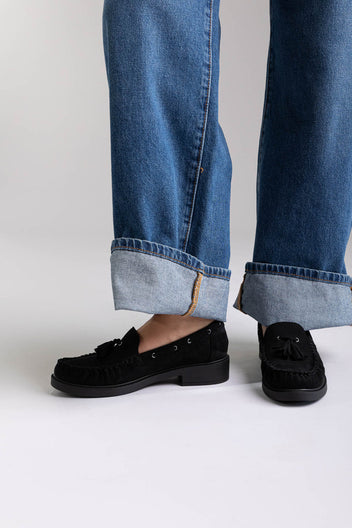 Black suede tassel loafers with flat sole, side view on a model in wide-leg jeans