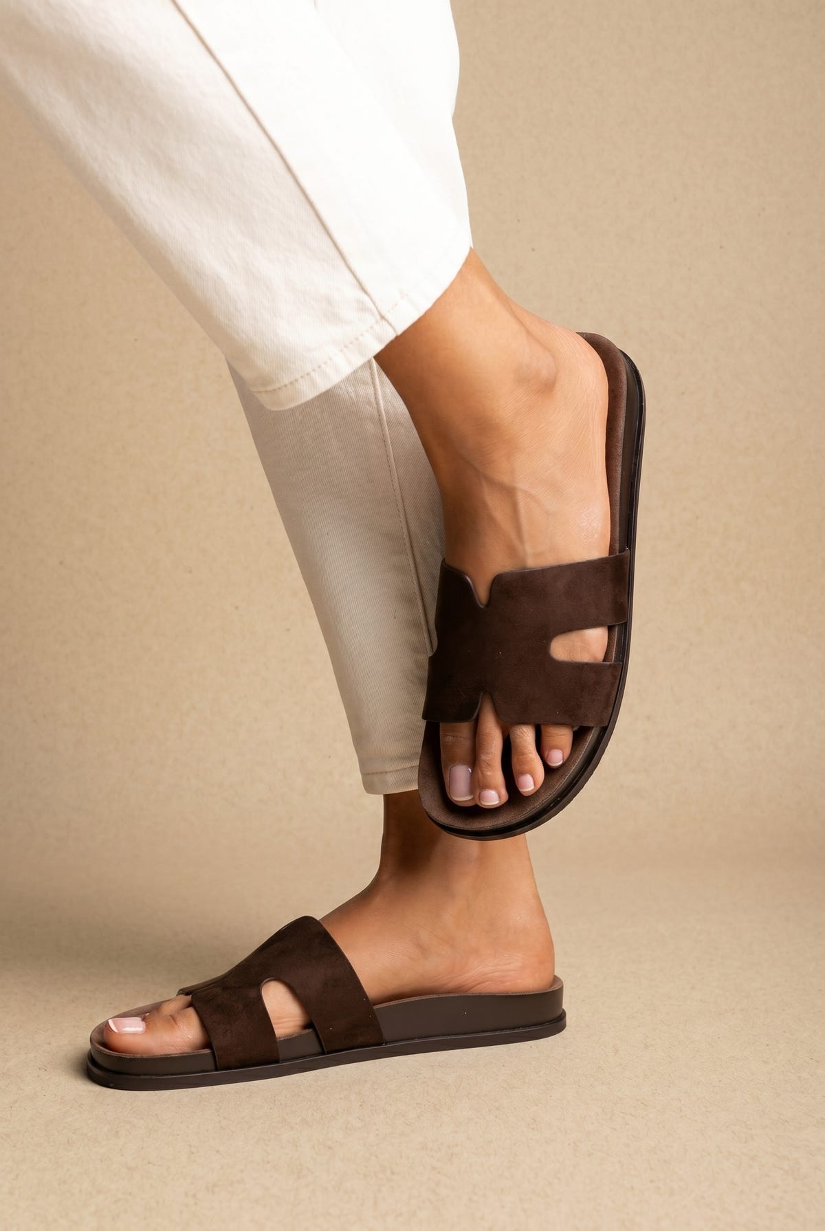 Dark brown suede H-band cut-out slide sandals on a flat footbed, worn on a model with cream trousers, side view
