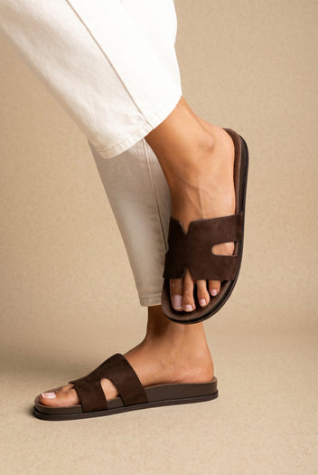 Dark brown suede H-band cut-out slide sandals on a flat footbed, worn on a model with cream trousers, side view