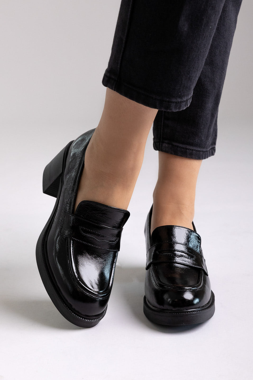 Black patent leather penny loafers with block heel and chunky sole, angled view on a model in black jeans