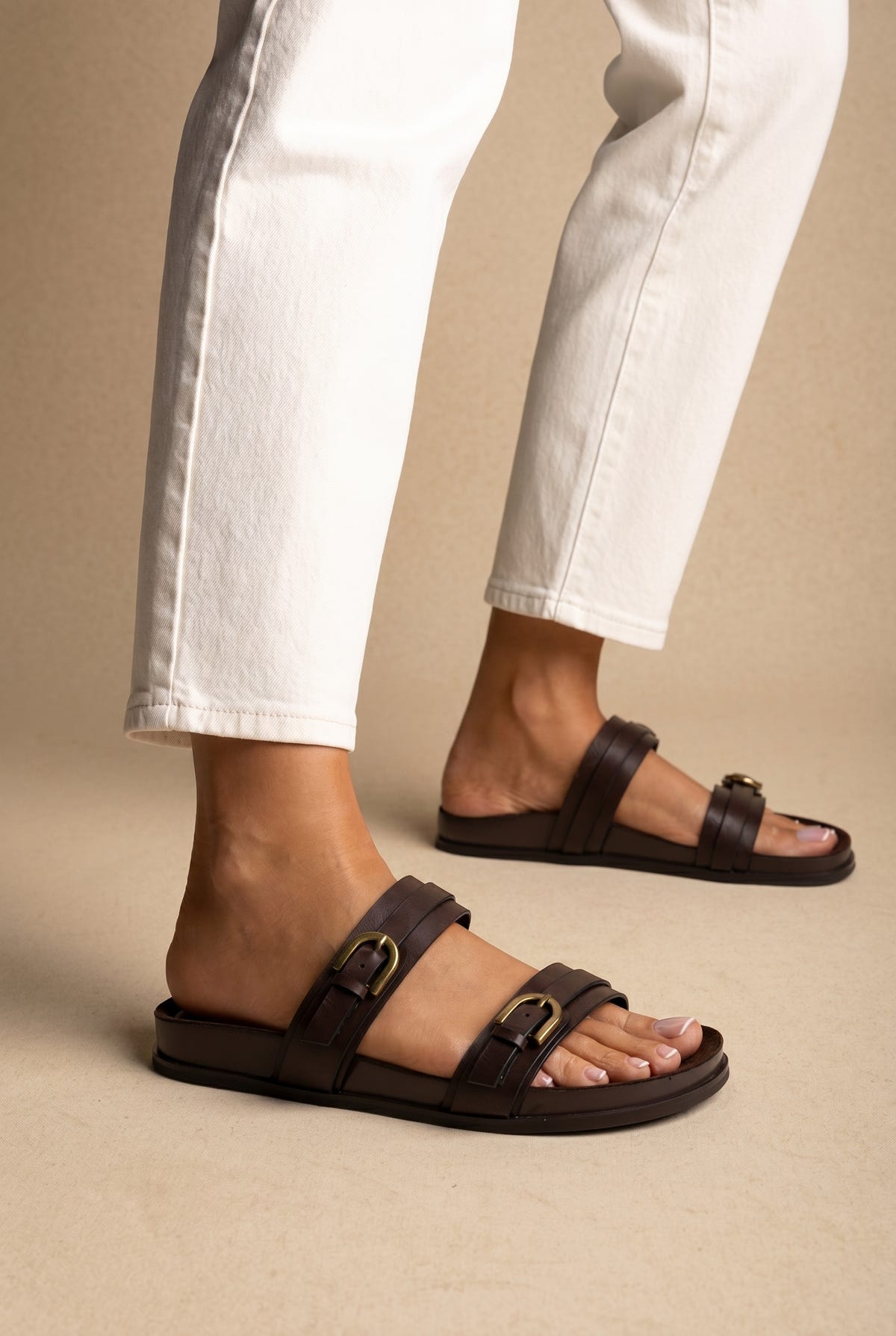 Dark brown leather double-strap slide sandals with gold buckles on a flat footbed, three-quarter view worn on a model