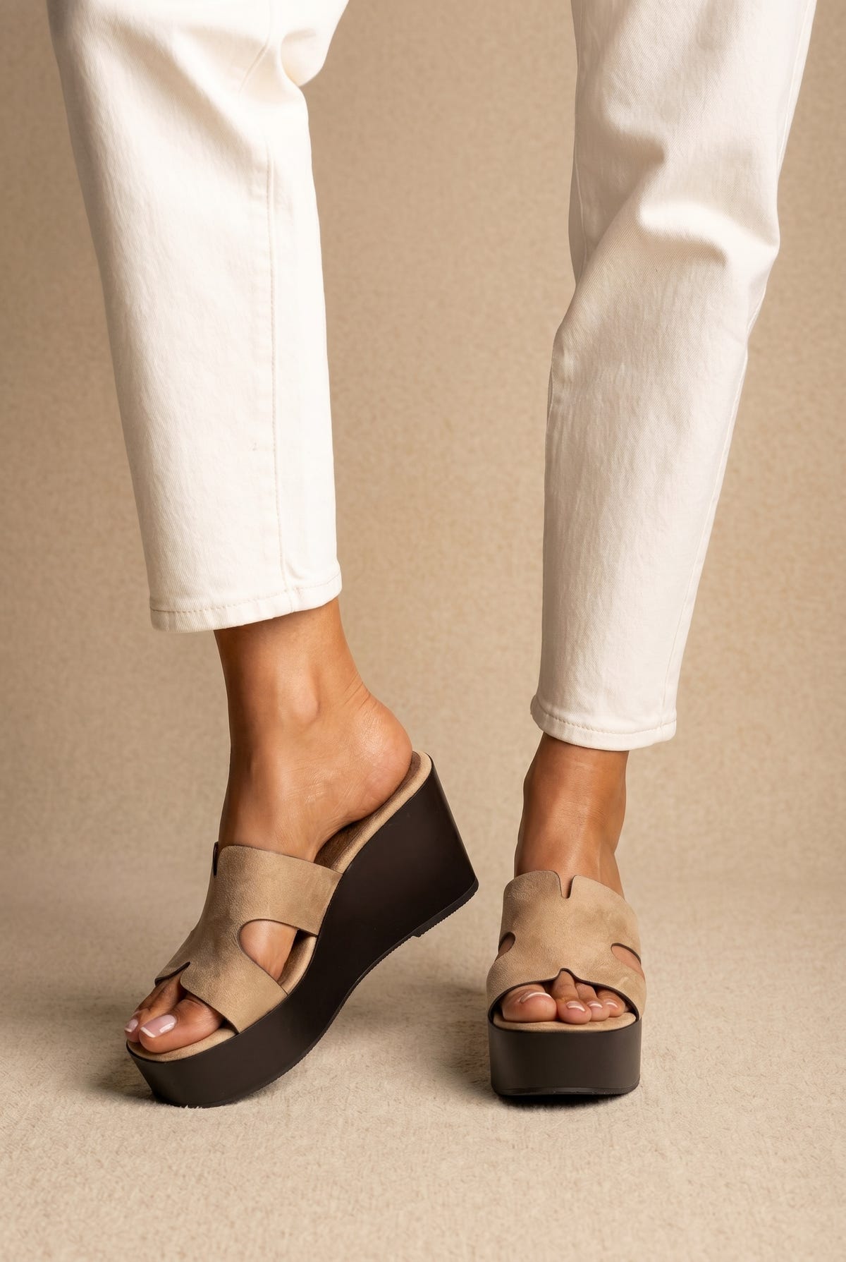 Beige suede platform wedge slides with a cut-out strap and black chunky sole, front view worn on a model with white jeans