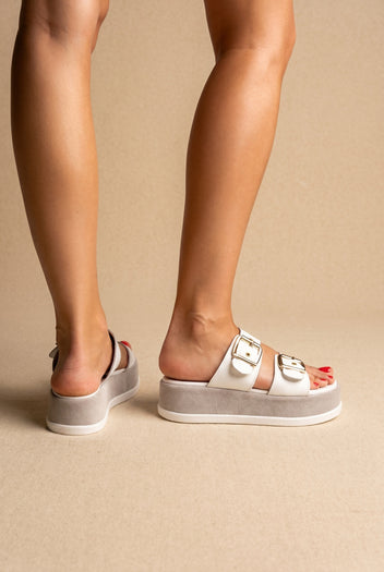 White leather platform slides with two gold buckle straps and grey suede platform with white edge, side view on a model