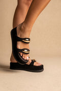 Close-up of black suede strap platform slides with shiny gold oval ornaments on a chunky sole, worn on a model