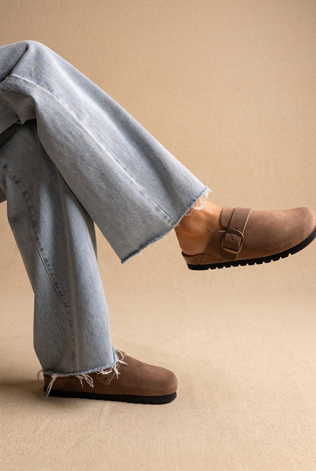Taupe suede clogs with buckle strap and black ridged sole, side view worn with wide-leg jeans on a model
