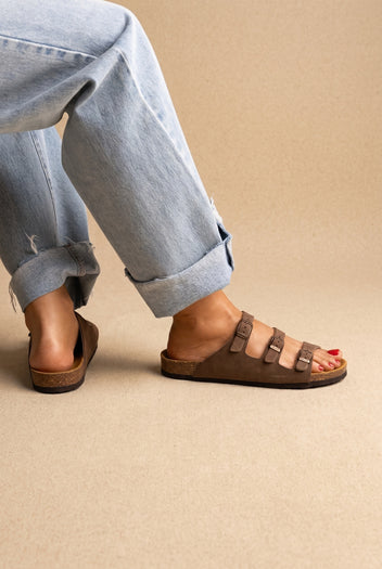Brown nubuck three-strap buckle sandals with cork footbed and black sole, side view worn with jeans on a model