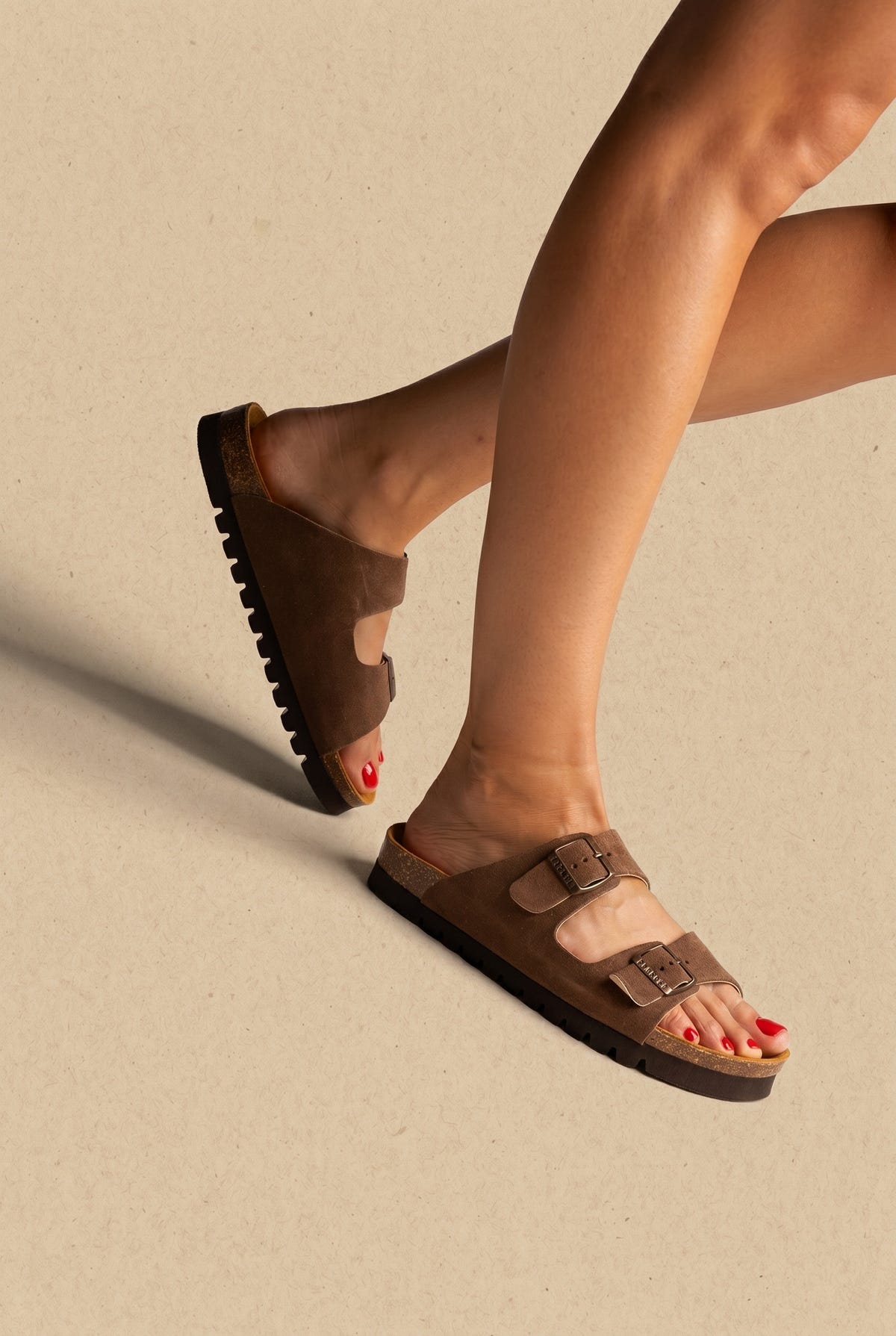 Brown suede two-strap buckle sandals with cork footbed and black lugged sole, worn on a model, side view