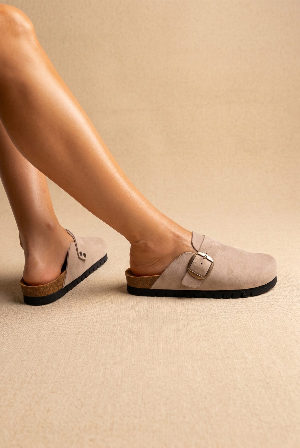 Light taupe suede clogs with a gold buckle strap, cork footbed edge and black sole, worn on a model, side view