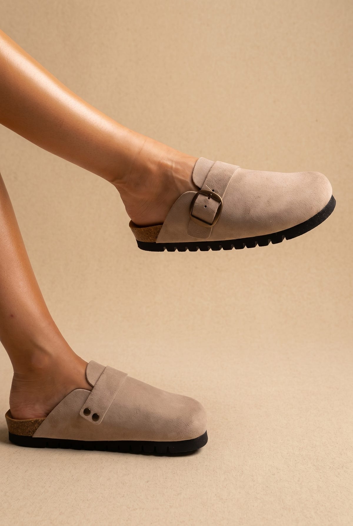 Beige suede clogs with a gold buckle strap and chunky black lugged sole, worn on a model, three-quarter view