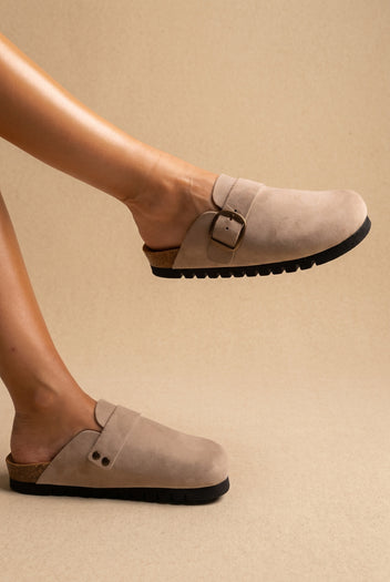 Beige suede clogs with a gold buckle strap and chunky black lugged sole, worn on a model, three-quarter view