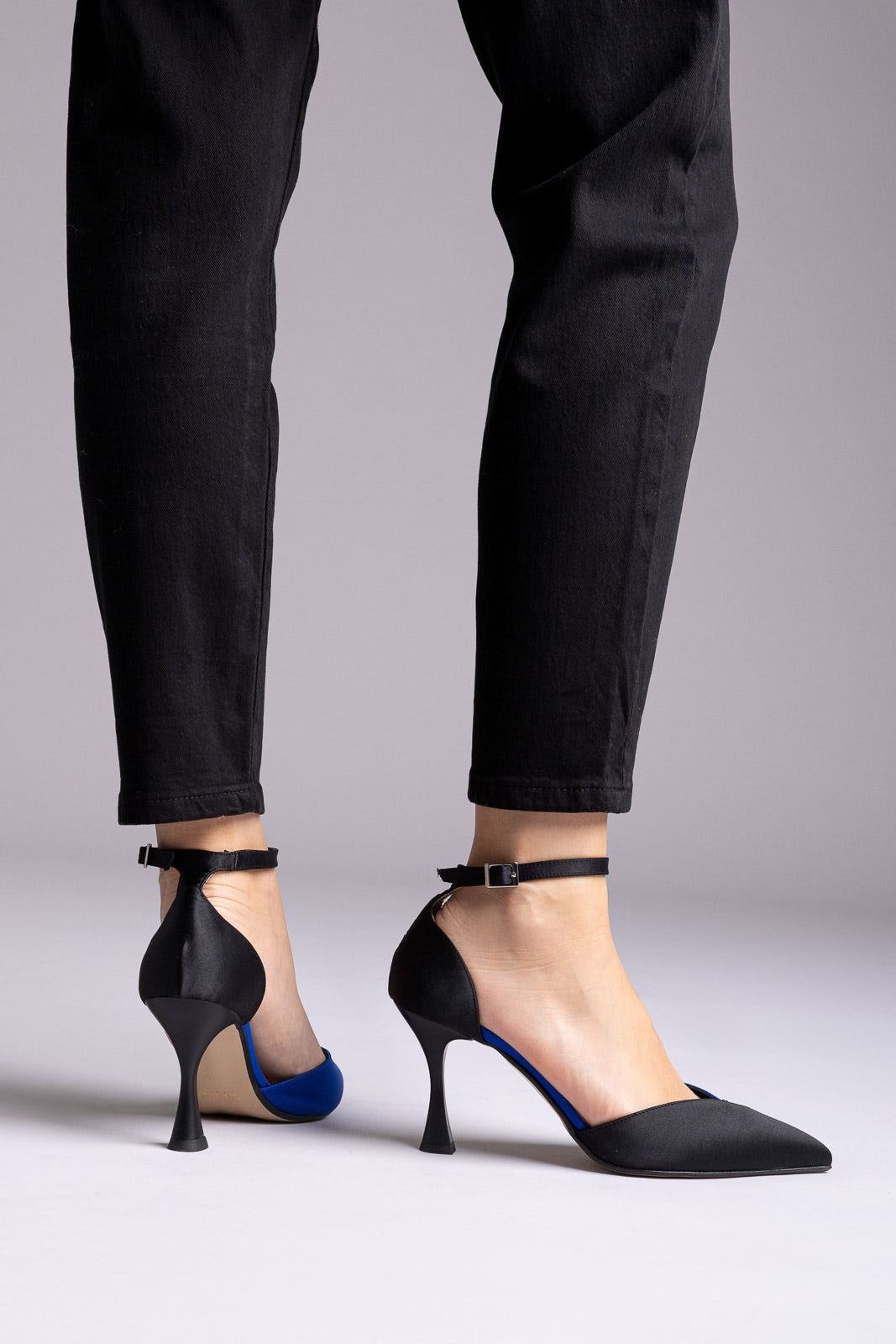 Black satin ankle-strap pumps with blue accent at the inner arch and flared heel, rear view with black jeans