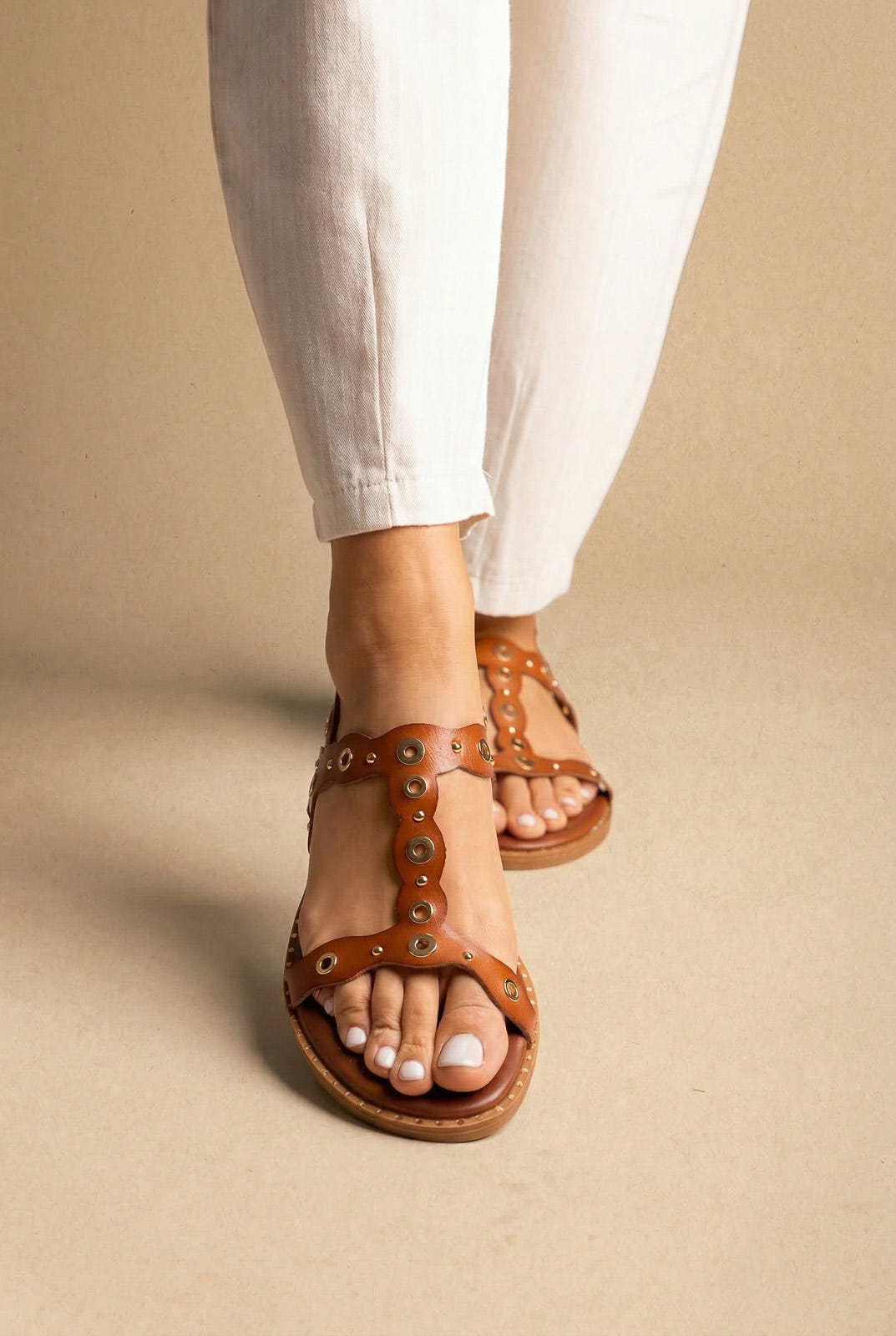 Tan leather flat sandal with T-strap front and round metal eyelet studs, worn with white linen trousers