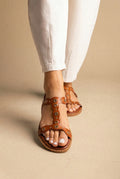 Tan leather flat sandal with T-strap front and round metal eyelet studs, worn with white linen trousers