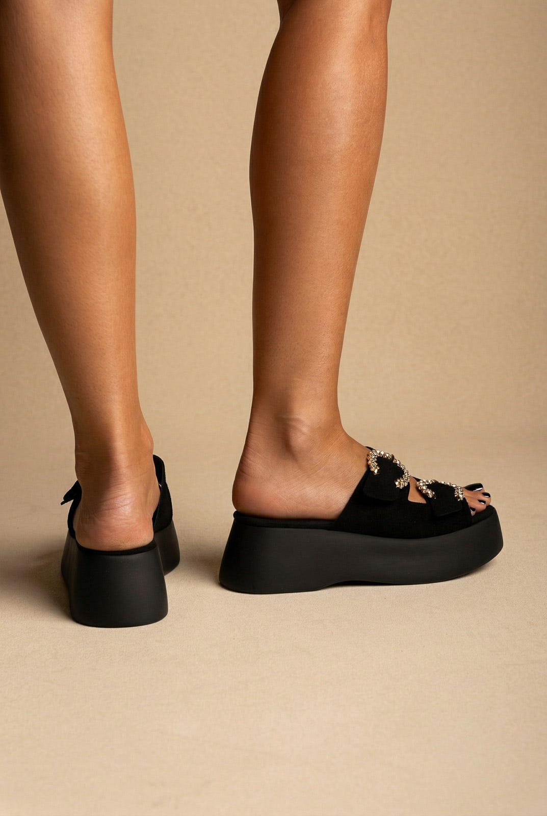 Black suede platform slides with two crystal-trimmed buckle straps and a thick sole, back view on a model