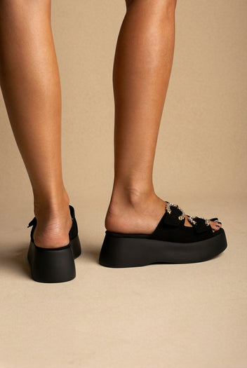 Black suede platform slides with two crystal-trimmed buckle straps and a thick sole, back view on a model
