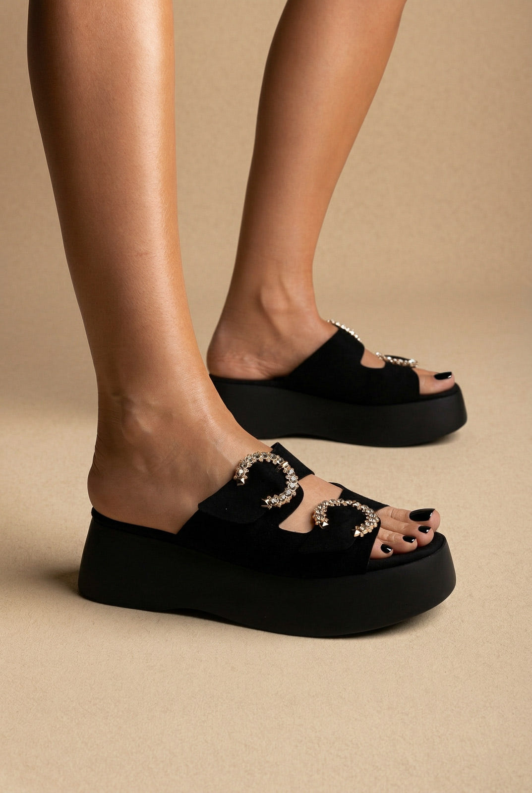 Black suede platform slides with two crystal-trimmed round buckles and a chunky sole, side view on a model
