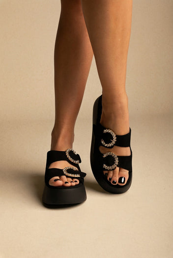 Black suede platform slides with two straps and crystal-trimmed round buckles, front view on a model