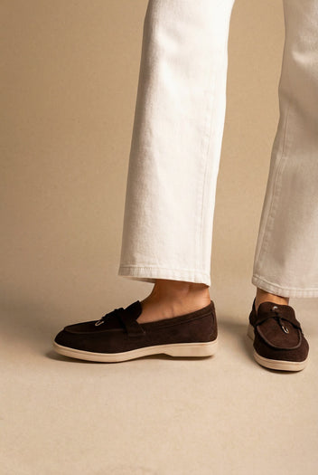 Side view of dark brown suede loafers with cream soles and tie detail, worn with white jeans