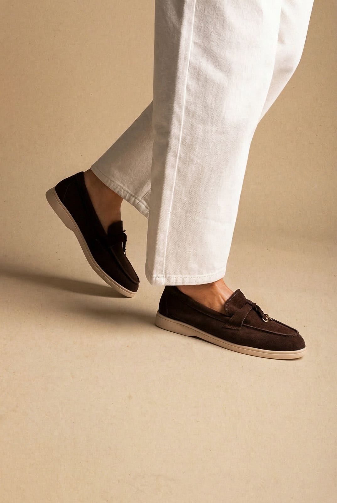 Model in dark brown suede loafers with knotted tie trim and cream sole, one foot raised