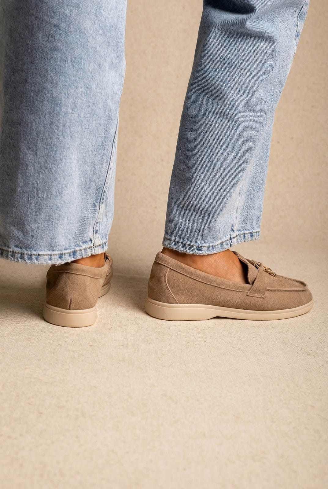 Back view of taupe suede loafers with cream soles and small metal tie detail, worn with light-wash jeans