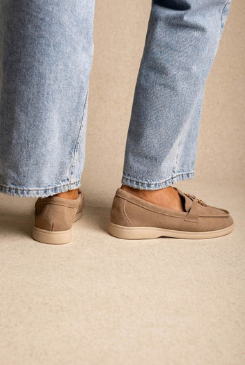 Back view of taupe suede loafers with cream soles and small metal tie detail, worn with light-wash jeans