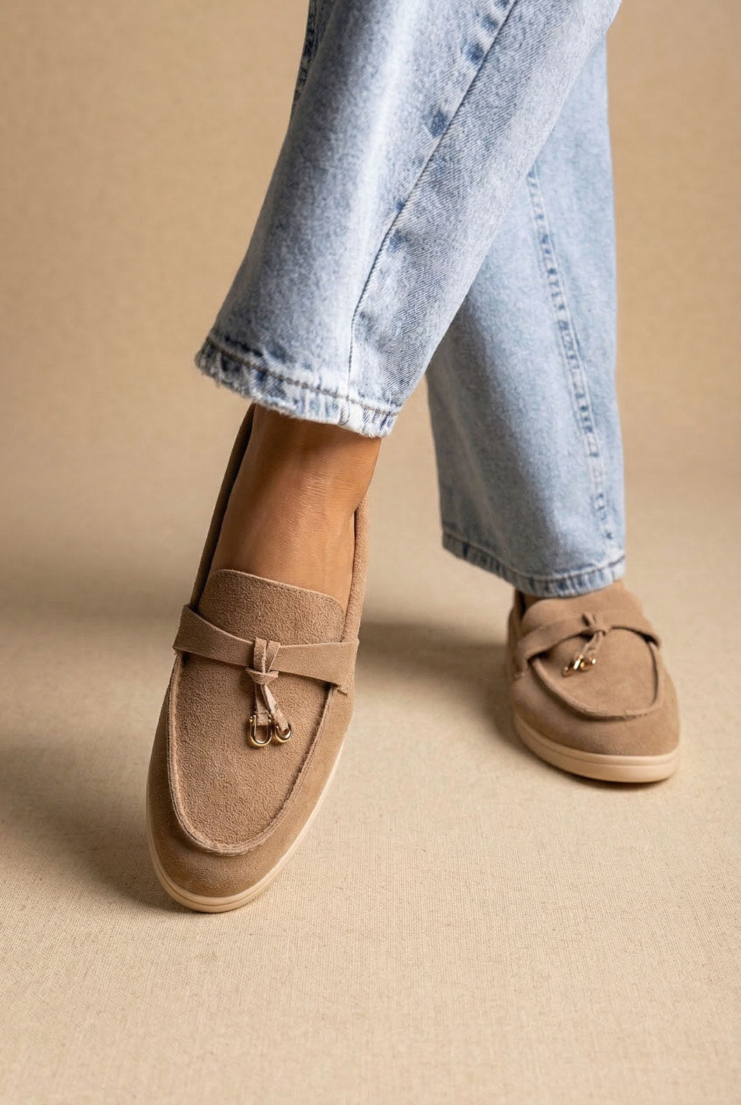 Taupe suede slip-on loafer with a leather strap and gold-tone tassel charm, cream sole, worn with light blue jeans