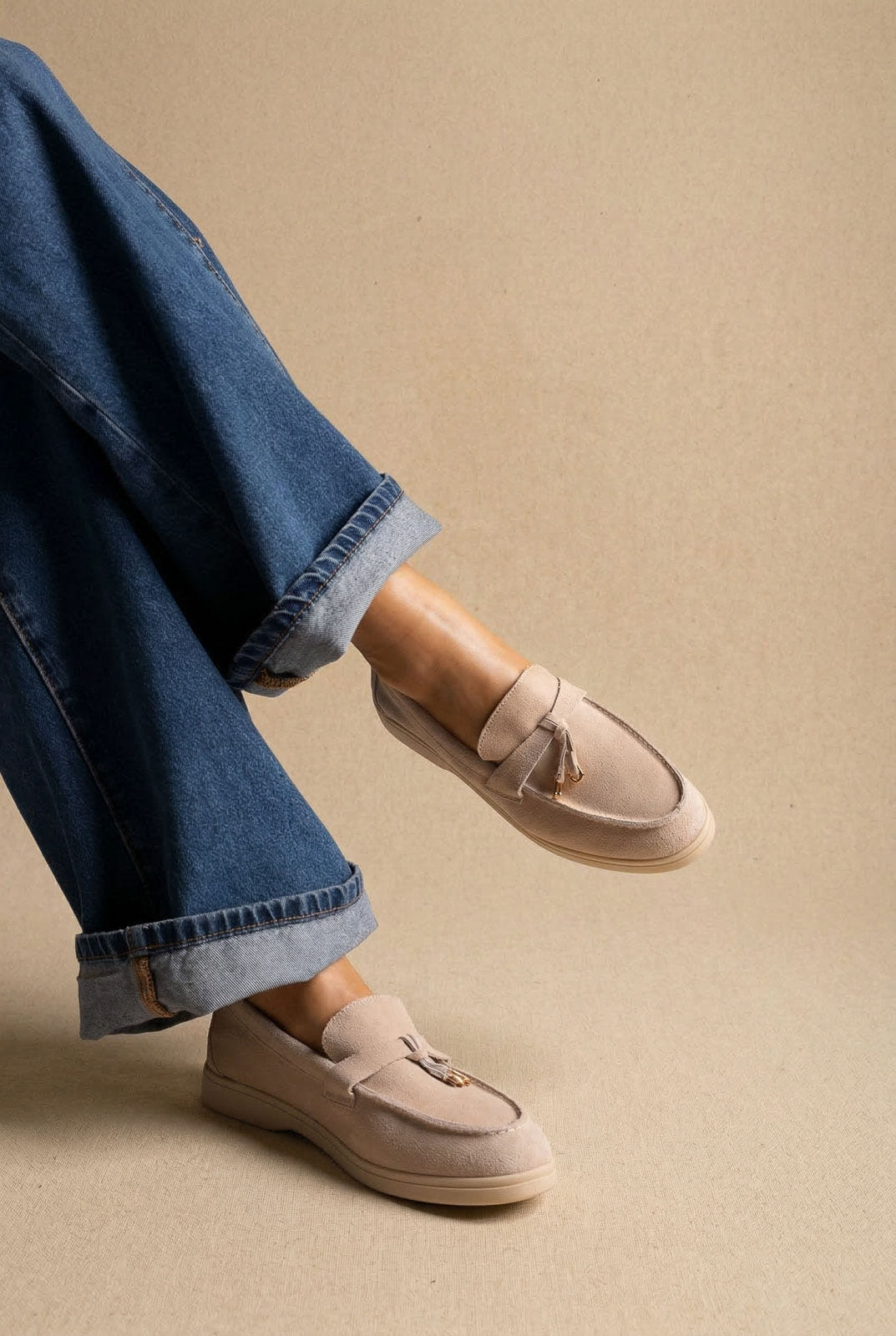 Pair of sand suede loafers with small tassel trim and light sole, worn with cuffed blue jeans in a crossed leg pose