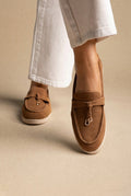 Close-up of tan suede loafers with knotted tie and gold-tone ring charm on cream soles