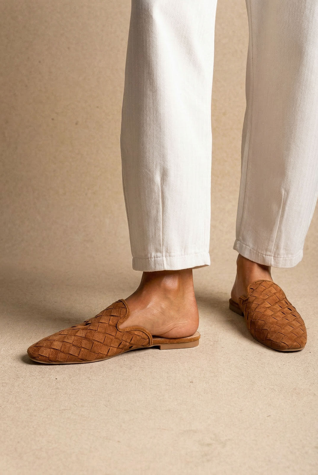 Model wearing tan suede woven mules with pointed toe and flat sole, paired with cream trousers