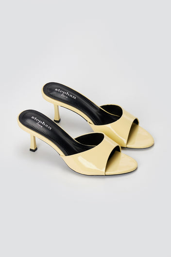 Pair of pale yellow patent leather heeled mules with black insoles and slim flared heels, three-quarter view