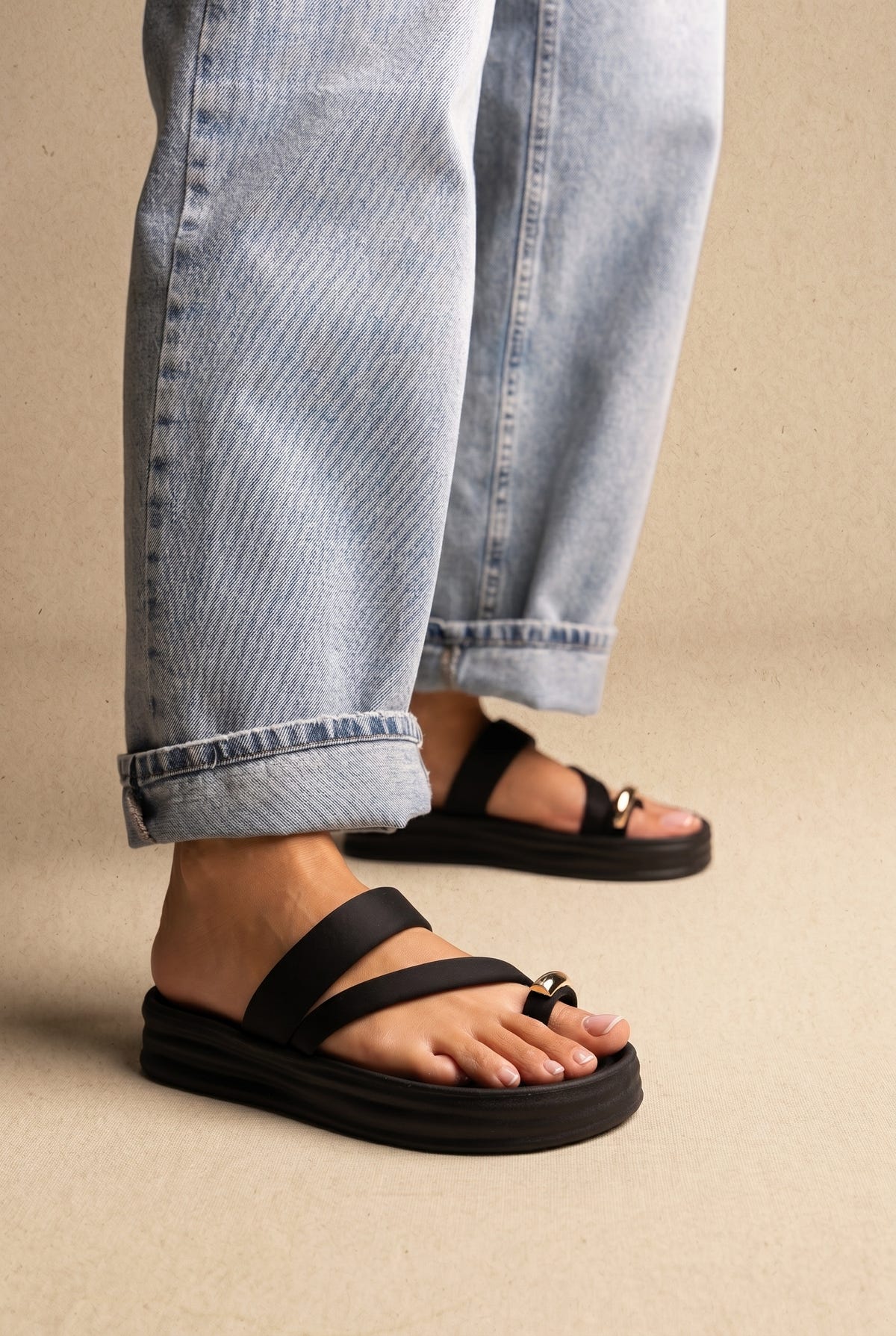 Black flatform slide sandals with two straps, toe loop and gold metal detail, worn on a model with light blue jeans, side view