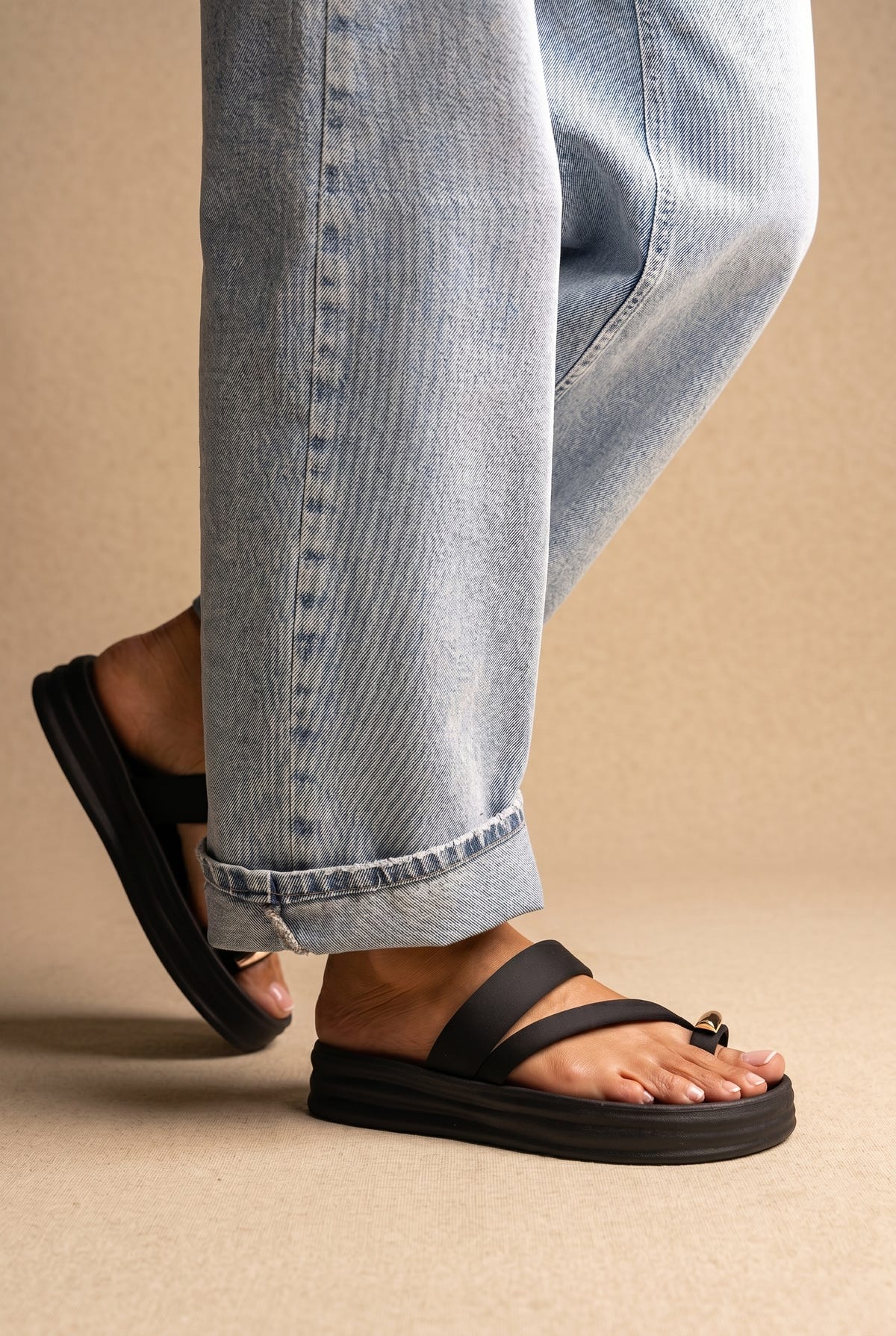 Black flatform sandals with toe loop, thin straps and gold detail on a thick sole, worn with jeans, side view