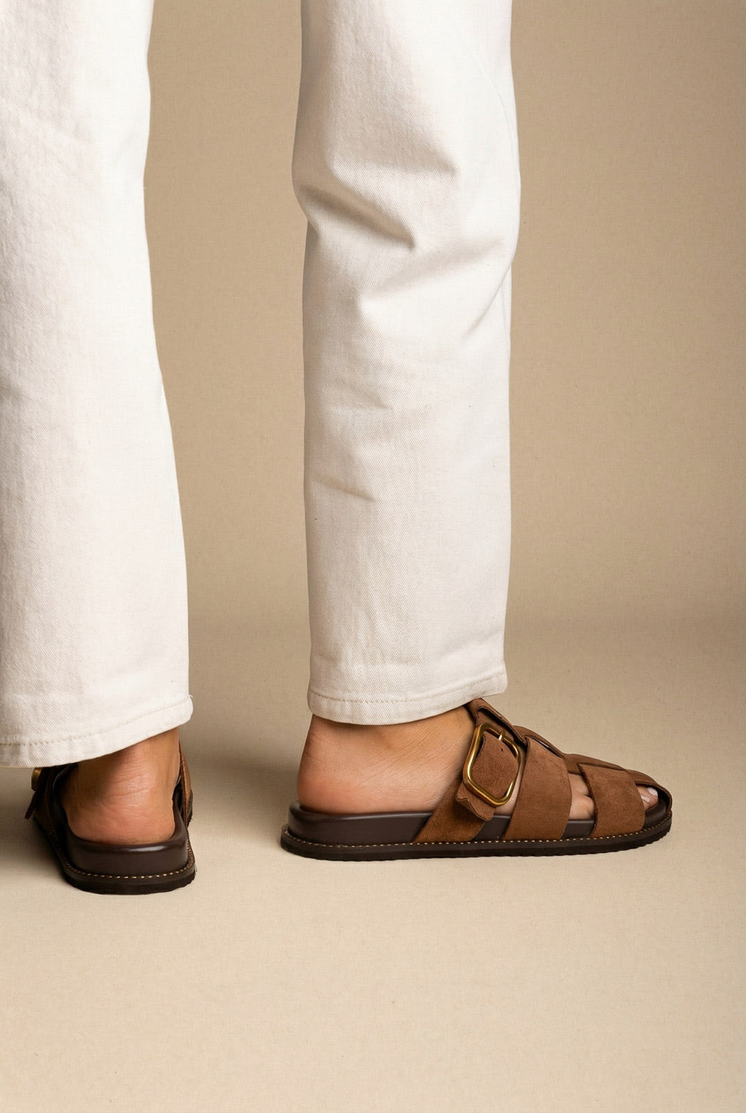 Brown suede cage-strap slides with a large gold buckle, worn with white jeans, back and side view on a model