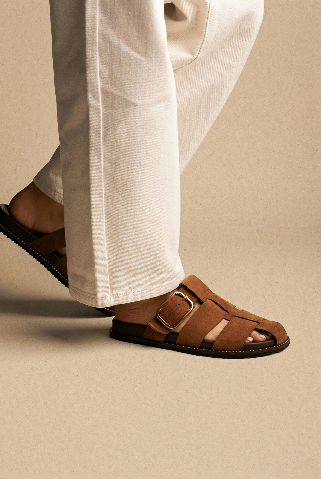 Brown suede closed-toe cage slides with a large gold buckle on a flat sole, worn with white jeans, side view