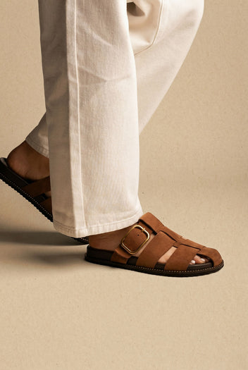 Brown suede closed-toe cage slides with a large gold buckle on a flat sole, worn with white jeans, side view