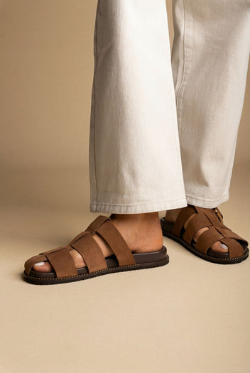 Brown suede fisherman slide sandals with cage straps and a dark flat sole, worn with white wide-leg jeans on a model