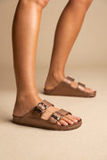 Copper bronze metallic molded two-strap slide sandals with gold-tone buckles, worn on a model, side three-quarter view