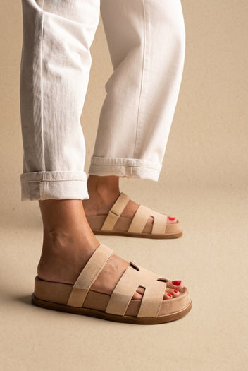 Beige suede chunky slide sandals with wide cut-out straps and a thick sole, side view on a model in white jeans