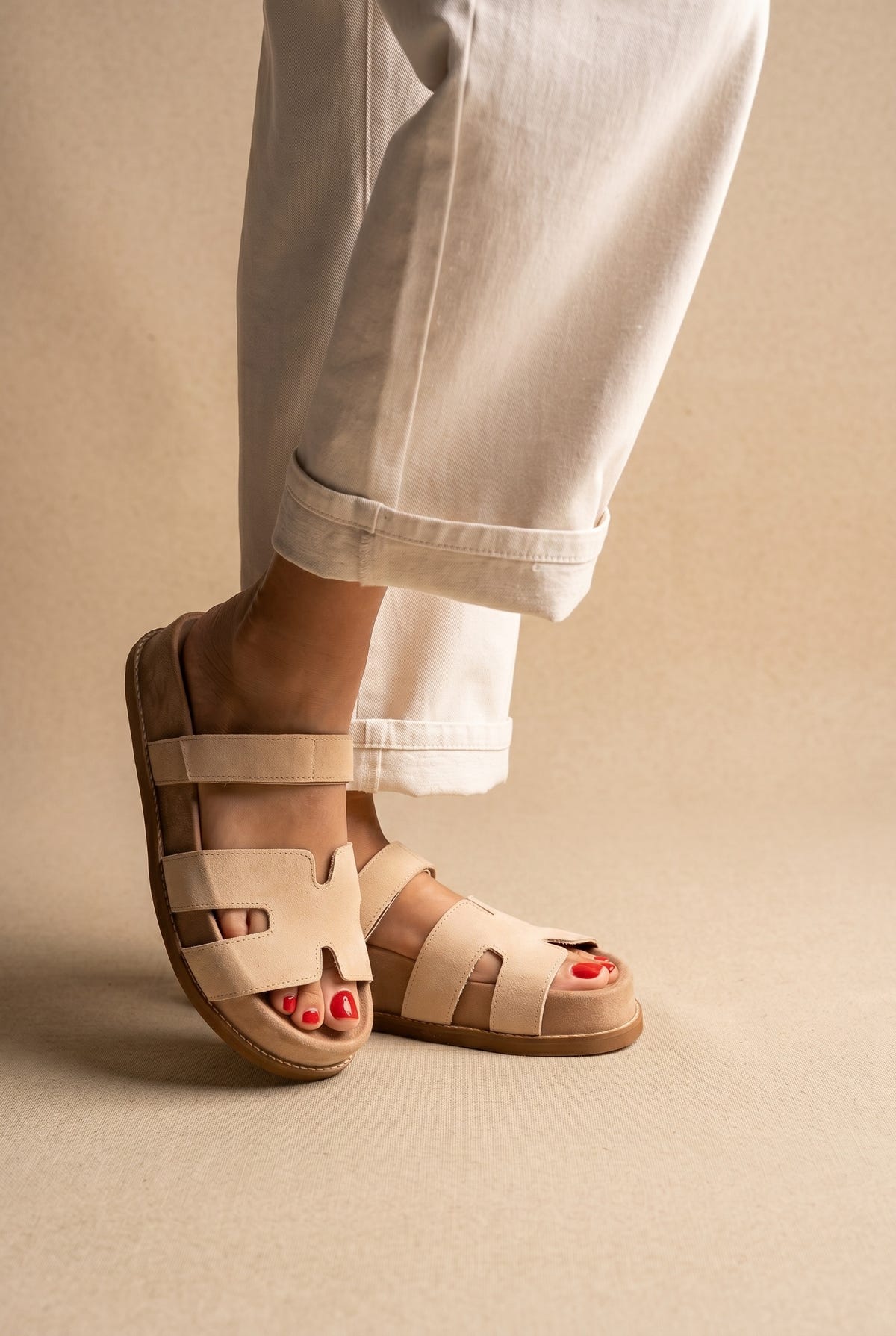 Beige suede chunky slide sandals with wide cut-out straps and a back strap, three-quarter view on a model in white jeans