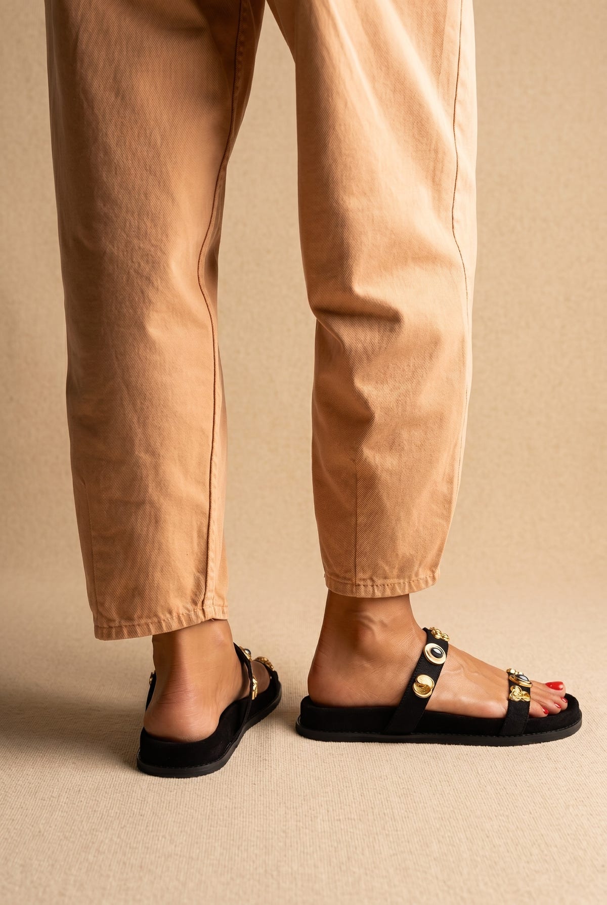 Back and side view of black suede slide sandals with two straps and gold embellishments, worn with tan trousers