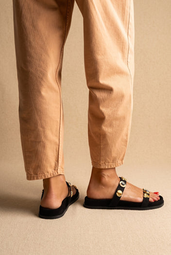 Back and side view of black suede slide sandals with two straps and gold embellishments, worn with tan trousers