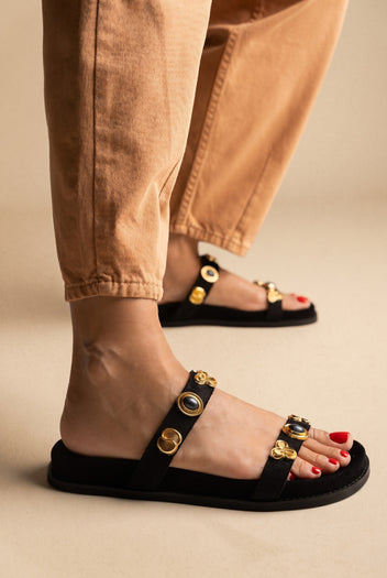 Black suede slide sandals with two straps and gold and blue stone embellishments on a thick flat sole, worn with tan trousers
